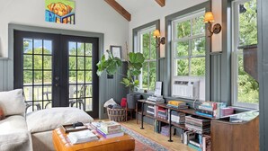 A cozy reading nook with a sofa, a coffee table with books, a bookshelf, and large windows with a view of greenery. - Farmhouse at Leeds by Avantstay Walled Garden + Soaking Tub + Private Barn Near Hudson (Leeds)