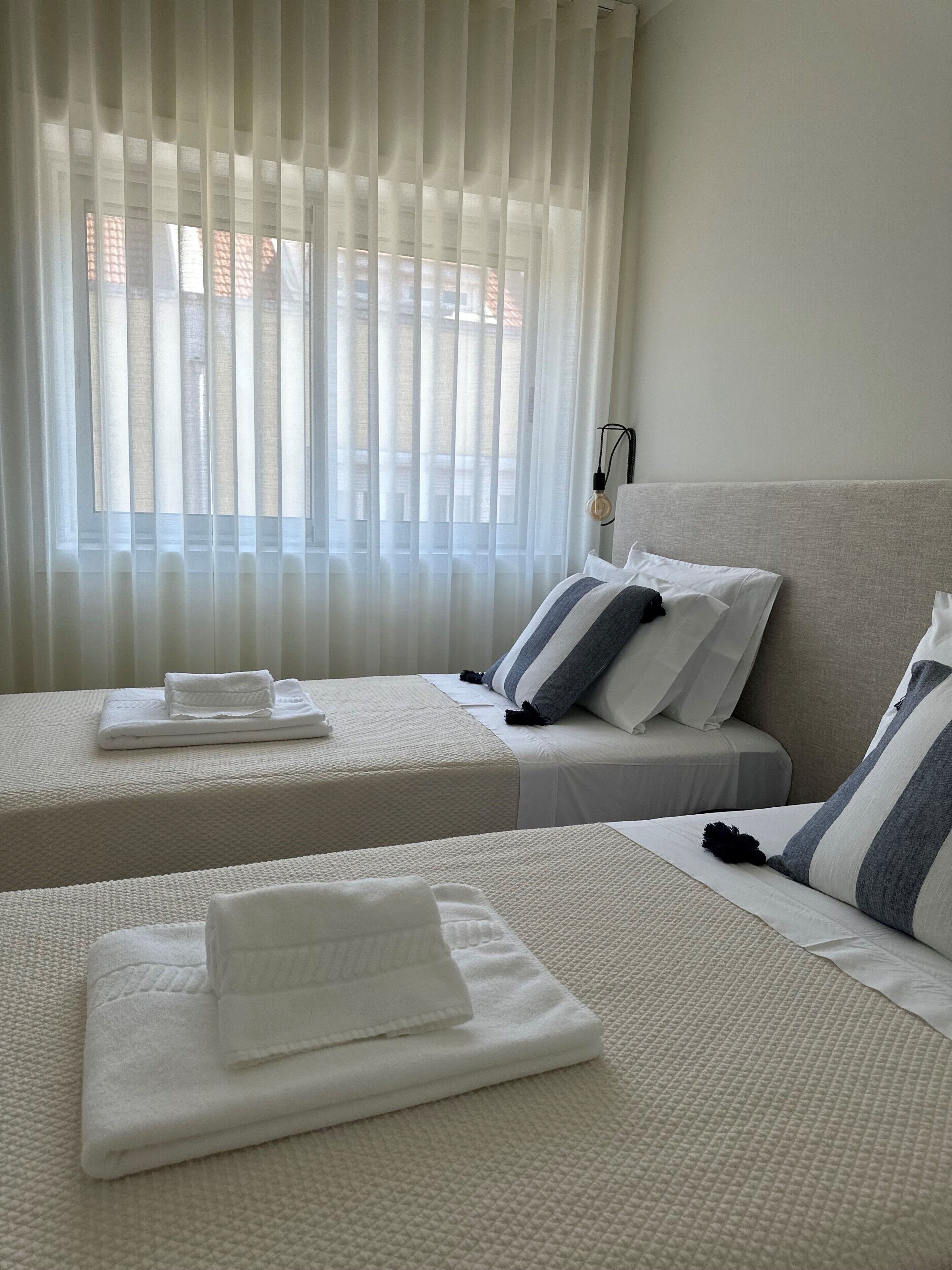 Camera d'albergo con due letti, biancheria bianca, una lampada e vista attraverso tende leggere.