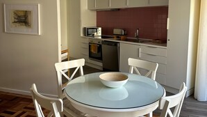 Een eethoek met een ronde glazen tafel, witte stoelen met beklede zitplaatsen, een wit kastje, een magnetron, een broodrooster en een betegelde spatwand in de keuken.