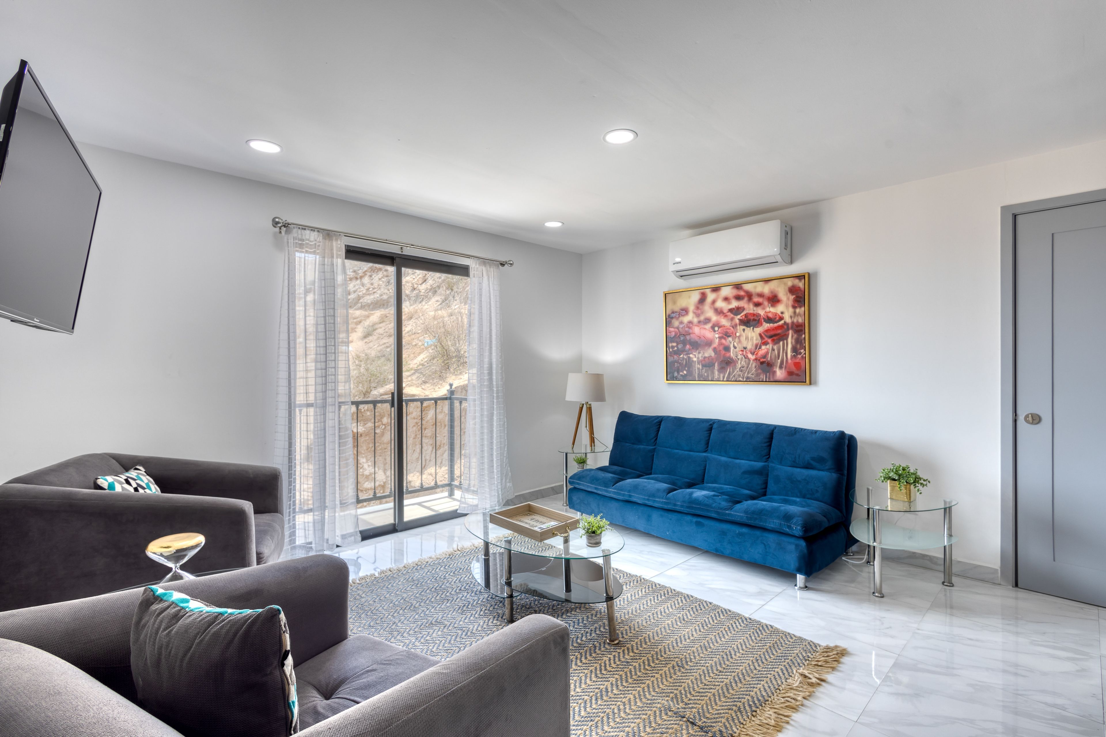 A modern living room with a grey sofa, a blue sofa, a glass coffee table, and a large window with curtains.