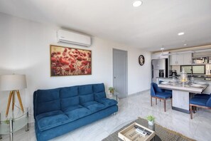 A modern living room with a blue sofa, a dining table with chairs, a kitchen area, and an air conditioning unit. - Dog-friendly Hotel Suite With Ocean-view Rooftop Infinity Pool, Central AC & gym (Cabo San Lucas)