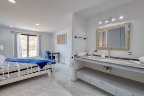 Un dormitori modern amb un llit, un escriptori, un mirall gran i un bany amb lavabo.