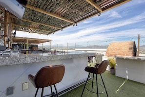 Uma área de bar ao ar livre com balcão de pedra, dois banquetas e vista para a cidade e montanhas.
