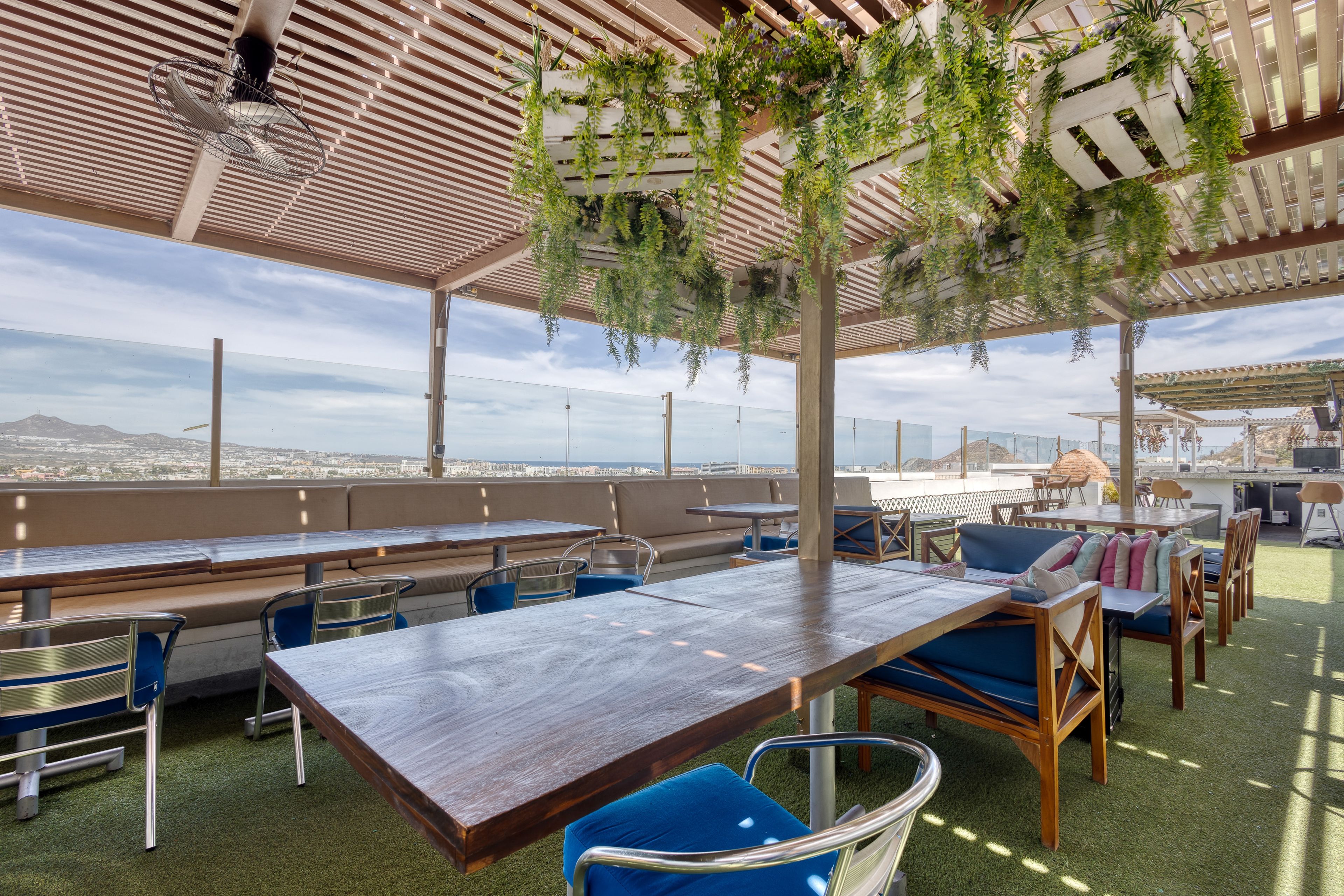 Un área de estar al aire libre con mesas y sillas, una pérgola con plantas colgantes y vistas de la ciudad.
