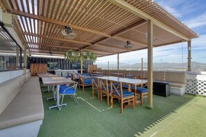 Uma área de estar externa com mesas e cadeiras, uma pérgula de madeira e vista para o horizonte da cidade.