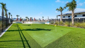 Een goed onderhouden kunstgrasgolfbaan met een heldere lucht, palmbomen en een zwembadgedeelte op de achtergrond.