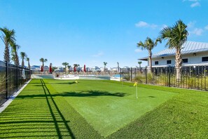 A well-maintained artificial grass golf course with a clear sky, palm trees, and a pool area in the background. - Sandy Cheeks | Pet-Friendly + Pool + Putting Green (Orange Beach)