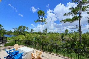 A patio with outdoor furniture, a table, and a view of a natural landscape with trees and a body of water. - Sandy Cheeks | Pet-Friendly + Pool + Putting Green (Orange Beach)
