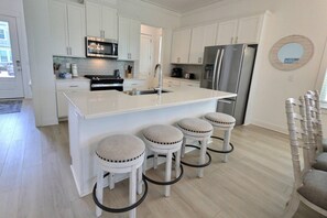 A modern kitchen with white cabinetry, a stainless steel refrigerator, a built-in oven, and a central island with bar stools. - Sandy Cheeks | Pet-Friendly + Pool + Putting Green (Orange Beach)