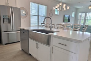 A modern kitchen with stainless steel appliances, a large island with a sink, white cabinetry, and a chandelier. - Sandy Cheeks | Pet-Friendly + Pool + Putting Green (Orange Beach)