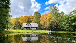 A lakeside cabin with a dock, surrounded by autumn foliage and a clear sky. - Private 13-Acre Retreat - Pond, Kayaks, Fire Pit & Trails. Pet-friendly! (Conneautville)