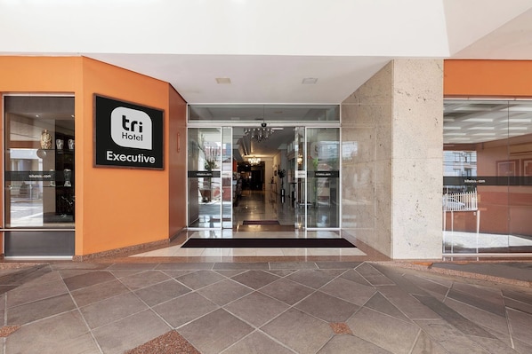 Entrada de un hotel con puerta de vidrio, un cartel que dice 'TRI Hotel Executive' y piso de baldosas.