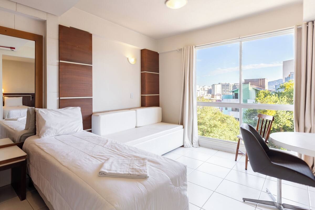 A hotel room with a bed, a desk, a chair, a large window with a city view, and a wall-mounted light.