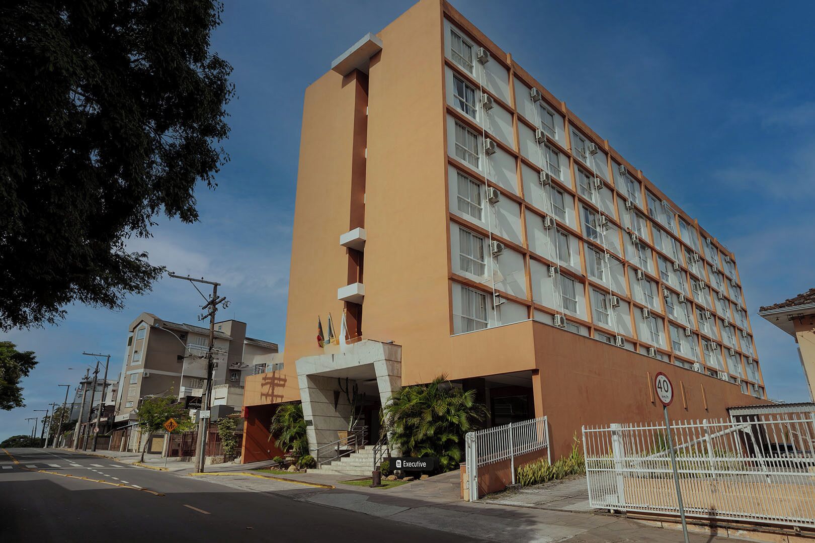 A multi-story hotel with a beige facade, a central entrance with steps, and a fenced parking area.