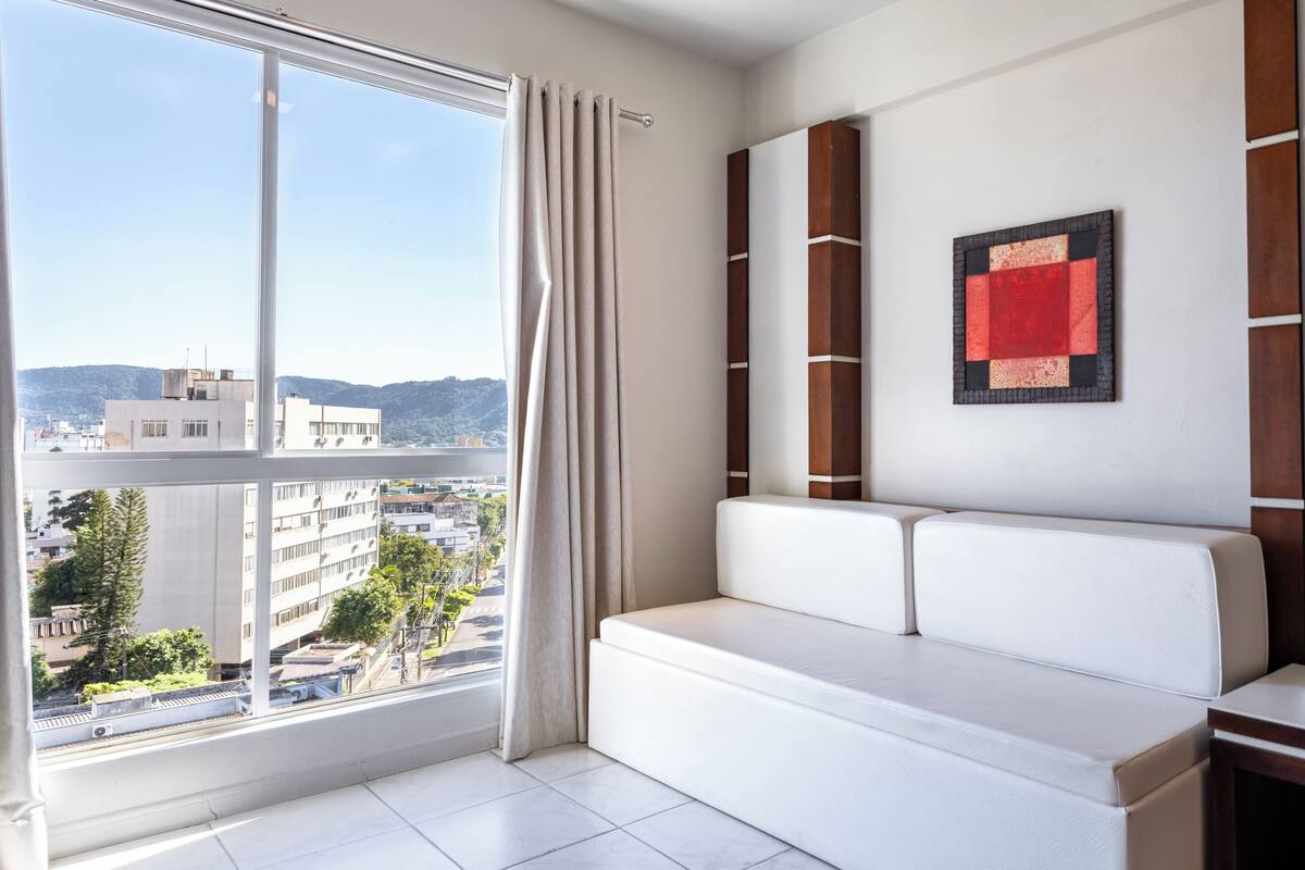 A modern room with a white sofa, a view of buildings through a large window, and a red and black framed picture on the wall.