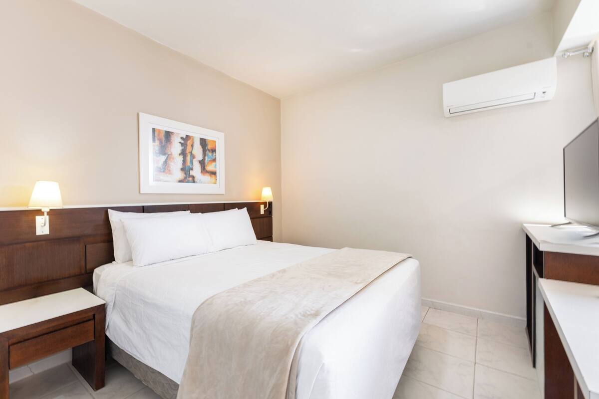 A hotel room with a bed, bedside tables, a desk, a chair, a TV, and an air conditioning unit.