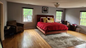 Un dormitorio con suelo de madera, una cama roja, dos ventanas, un ventilador de techo y un cuadro enmarcado en la pared.