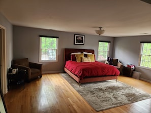 Ein Schlafzimmer mit Holzboden, einem roten Bett, zwei Fenstern, einem Deckenventilator und einem gerahmten Bild an der Wand.