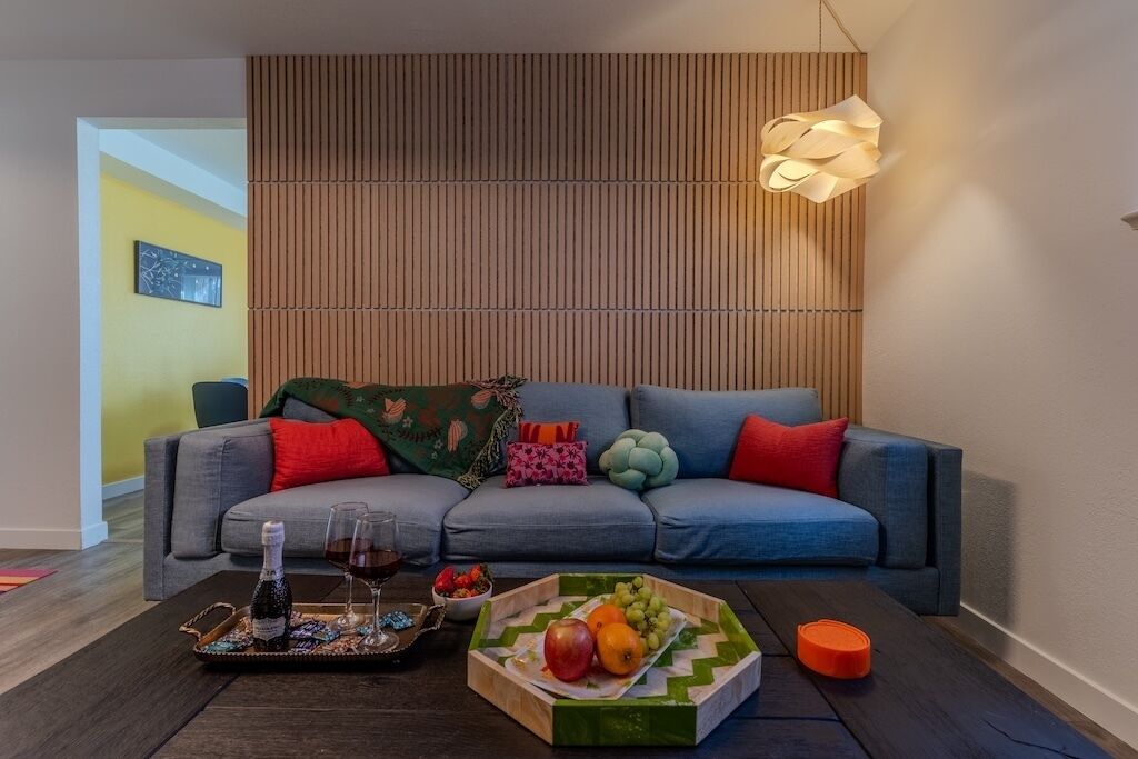 Ein modernes Wohnzimmer mit einem grauen Sofa, bunten Kissen, einer Holzpaneelwand und einer einzigartigen Pendelleuchte.