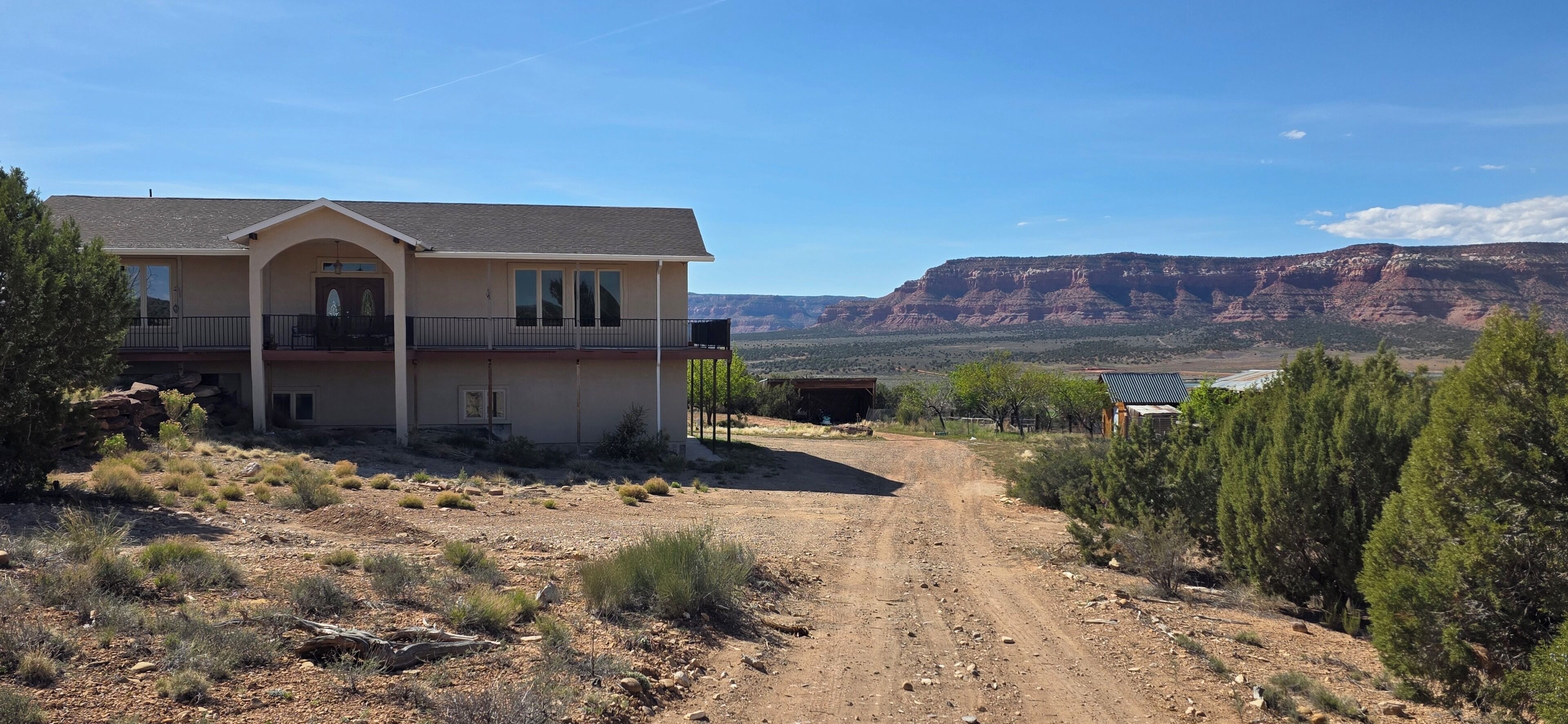 Una casa de dos pisos con balcón, camino de tierra y paisaje montañoso al fondo.