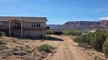 Una casa de dos pisos con balcón, camino de tierra y paisaje montañoso al fondo.
