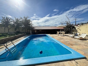A rectangular swimming pool with a ladder on one side, surrounded by a patio area with lounge chairs and a table. - Recanto Renascer (Aquiraz)