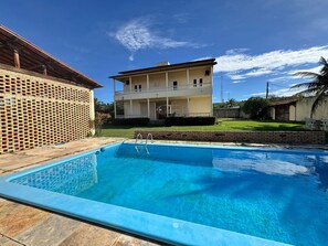 Una piscina con agua cristalina, una casa de dos pisos con balcón y un muro de ladrillo.