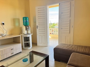 A room with a sofa, a glass-top coffee table, a water dispenser, a white cabinet, and a door leading to a balcony. - Recanto Renascer (Aquiraz)
