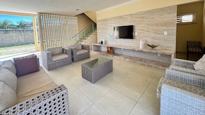 A spacious living area with a TV mounted on a stone wall, wicker furniture, and a staircase leading to an upper level. - Recanto Renascer (Aquiraz)