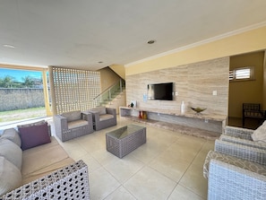 A spacious living area with a TV mounted on a stone wall, wicker furniture, and a staircase leading to an upper level. - Recanto Renascer (Aquiraz)