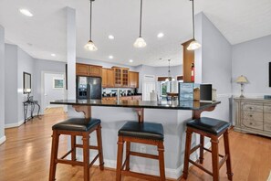 A kitchen with a central island, wooden bar stools, pendant lighting, and a view into the dining area.