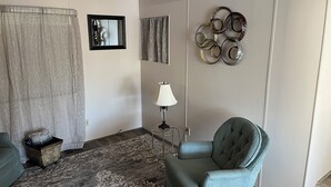 Un salón con un sillón verde, una alfombra estampada, una lámpara y decoración en la pared.