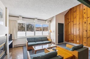 A room with a fireplace, a sofa, a coffee table, and a large window with a view of trees. - Timeshare Unit in Prestigious Stowe, VT (Stowe)