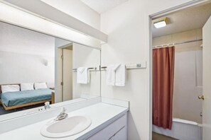 Une salle de bain d’hôtel avec un grand miroir, un lavabo blanc, une douche avec un rideau rouge et un lit recouvert d’une couverture bleue.