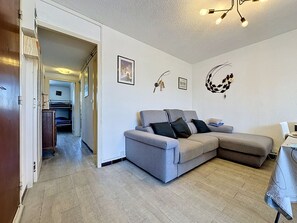 Een woonkamer met een grijze bank, een eettafel en een keuken die zichtbaar is door een open deur.