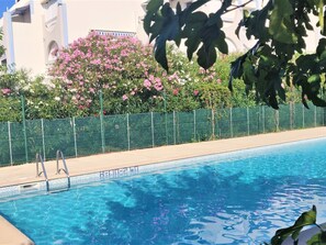 En swimmingpool med kristallklart blått vatten, omgiven av ett grönt staket och frodiga, rosa blommande växter.