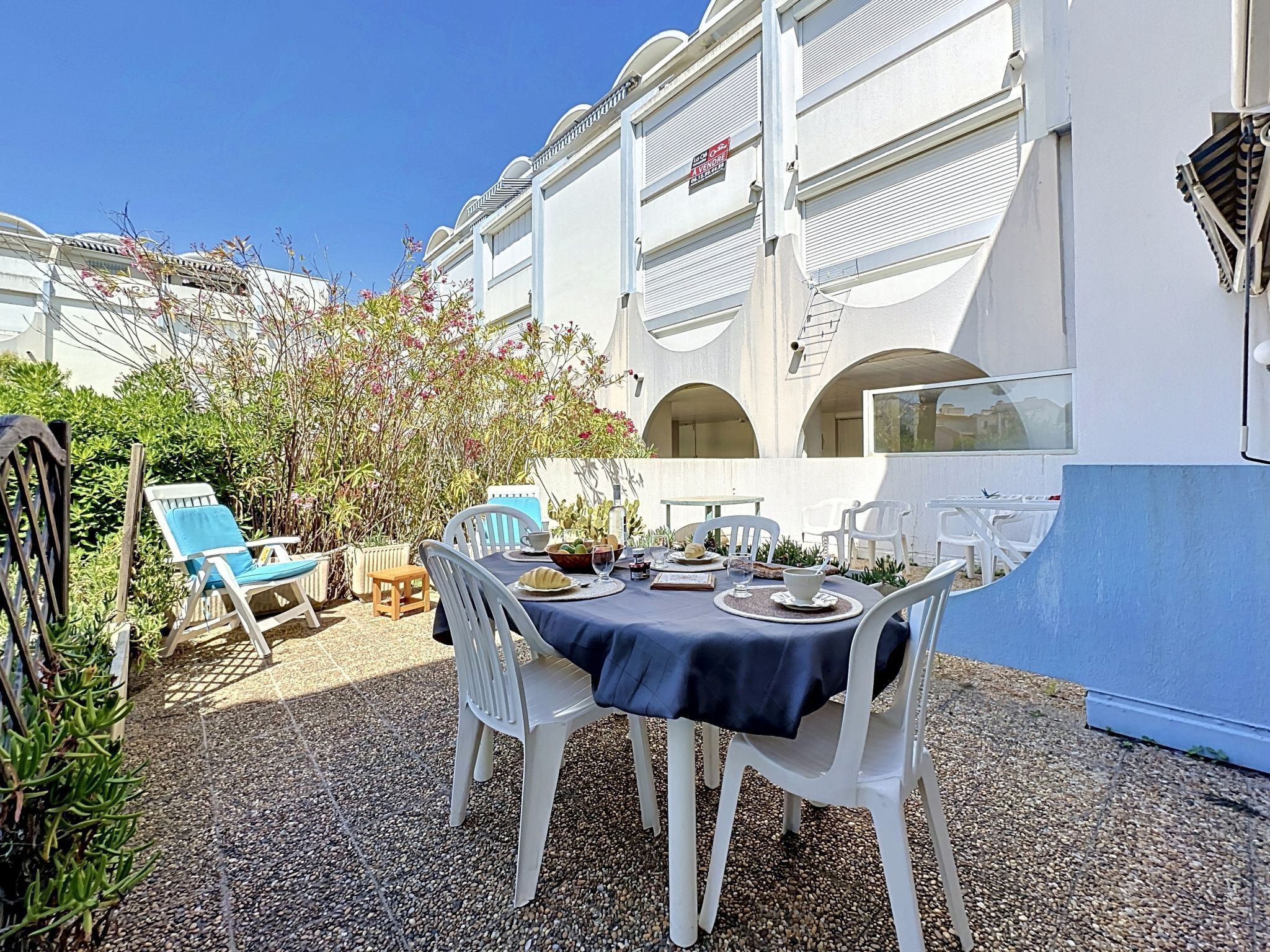 Un espace de restauration en plein air avec des chaises blanches et une table dressée pour deux, entouré de verdure et bordé d’un bâtiment doté de balcons.