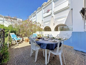 Un'area pranzo all'aperto con sedie bianche e un tavolo apparecchiato per due, immersa nel verde e affiancata da un edificio con balconi.