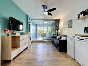 Ein modernes Wohnzimmer mit Flachbildfernseher, Deckenventilator, Sofa, Couchtisch und einer Küchenzeile inklusive Waschmaschine.