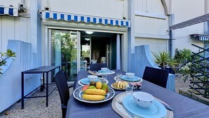 Ein Essbereich im Freien mit Tischen für zwei Personen, umgeben von Pflanzen und einem Gebäude mit einer blauen Markise.