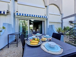 An outdoor dining area with tables set for two, surrounded by plants and a building with a blue awning. - Les Floralies II by Interhome (Le Grau du Roi)