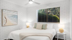 A bedroom with a white brick wall, a large window with blinds, a bed with a white bedspread, two bedside lamps, and a painting on the wall. - Southcoast Haven is the perfect spot for your next getaway (Shoalhaven Heads)