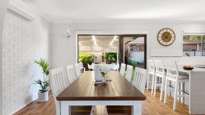 Ein Essbereich mit Holztisch und weißen Stühlen, einer Uhr an der Wand und Blick auf einen Garten durch eine offene Tür.