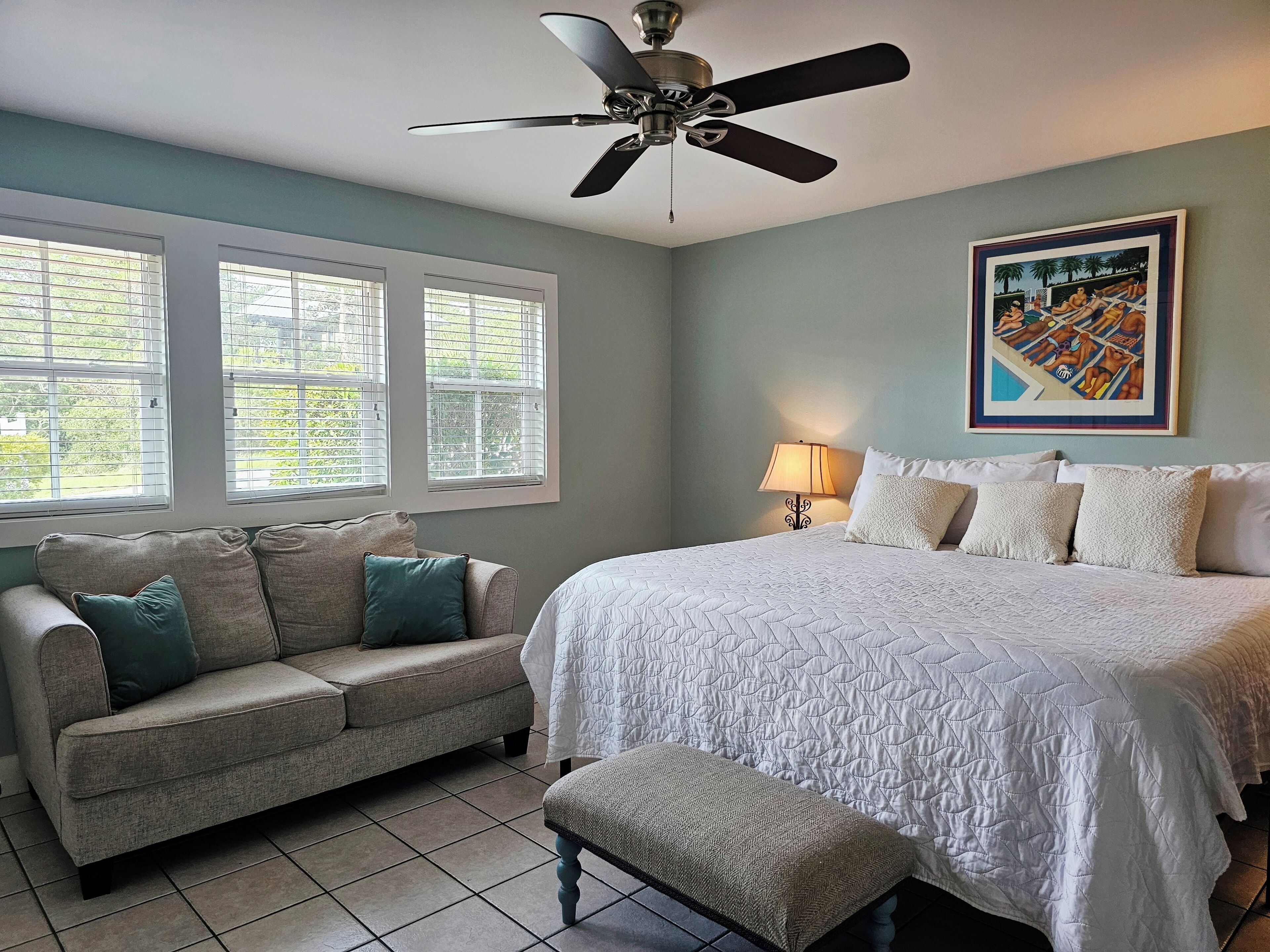 A bedroom with a bed, a sofa, a bench, a ceiling fan, and a window with blinds.
