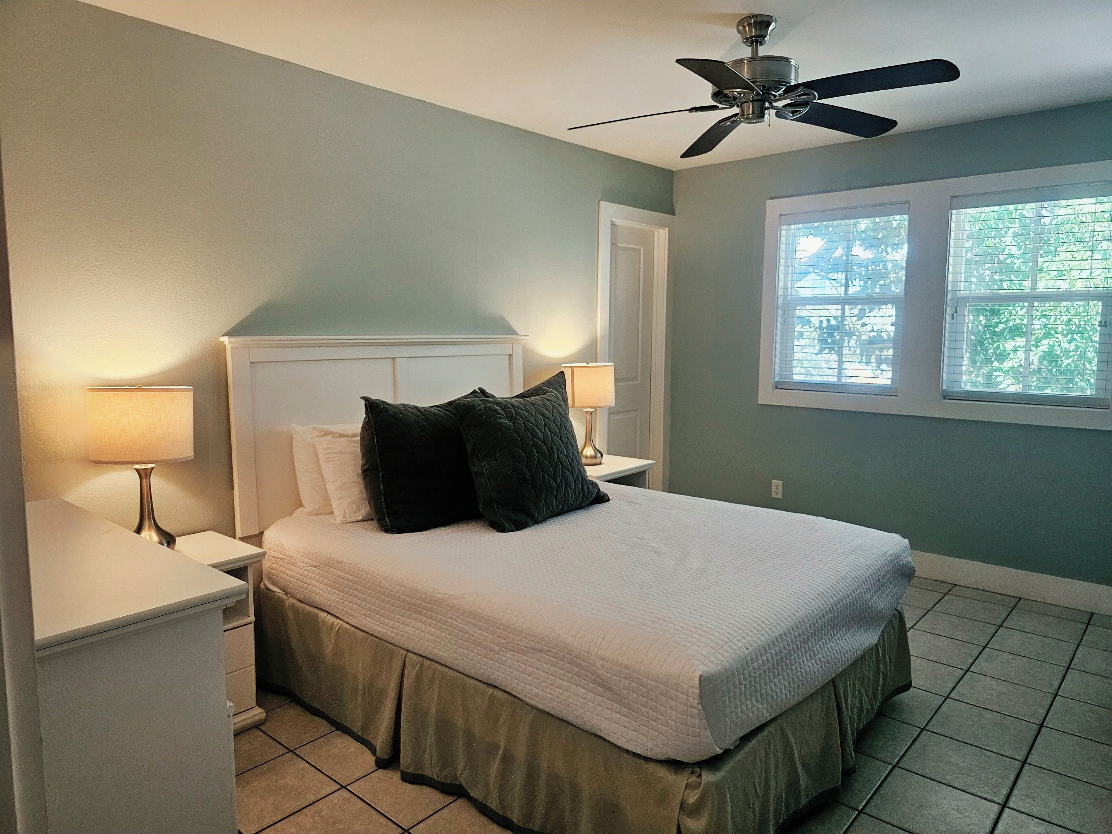 A bedroom with a bed, two bedside tables with lamps, a ceiling fan, a window with blinds, and a door.