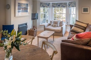 A cozy living room with a fireplace, comfortable seating, a table with a vase of flowers, and large windows with curtains. - The Retreat Stylish 4 bed  sea views   parking (Cowes)