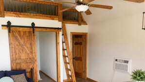 Quarto com cama, escrivaninha com banquinhos, ventilador de teto, vigas de madeira, escada e porta.
