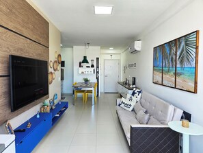A modern living room with a flat-screen TV, a blue entertainment unit, a dining area with yellow chairs, a kitchen with white cabinets, and a beach-themed wall art.