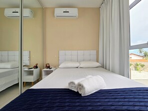 A hotel room with a bed, a nightstand, a mirror, an air conditioning unit, and a view of palm trees.