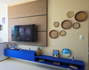 A modern living room with a flat-screen TV mounted on a wooden panel, a blue entertainment unit with decorative items, and a wall clock. - Apartamento 2 quartos VistaMar (Barra de São Miguel)